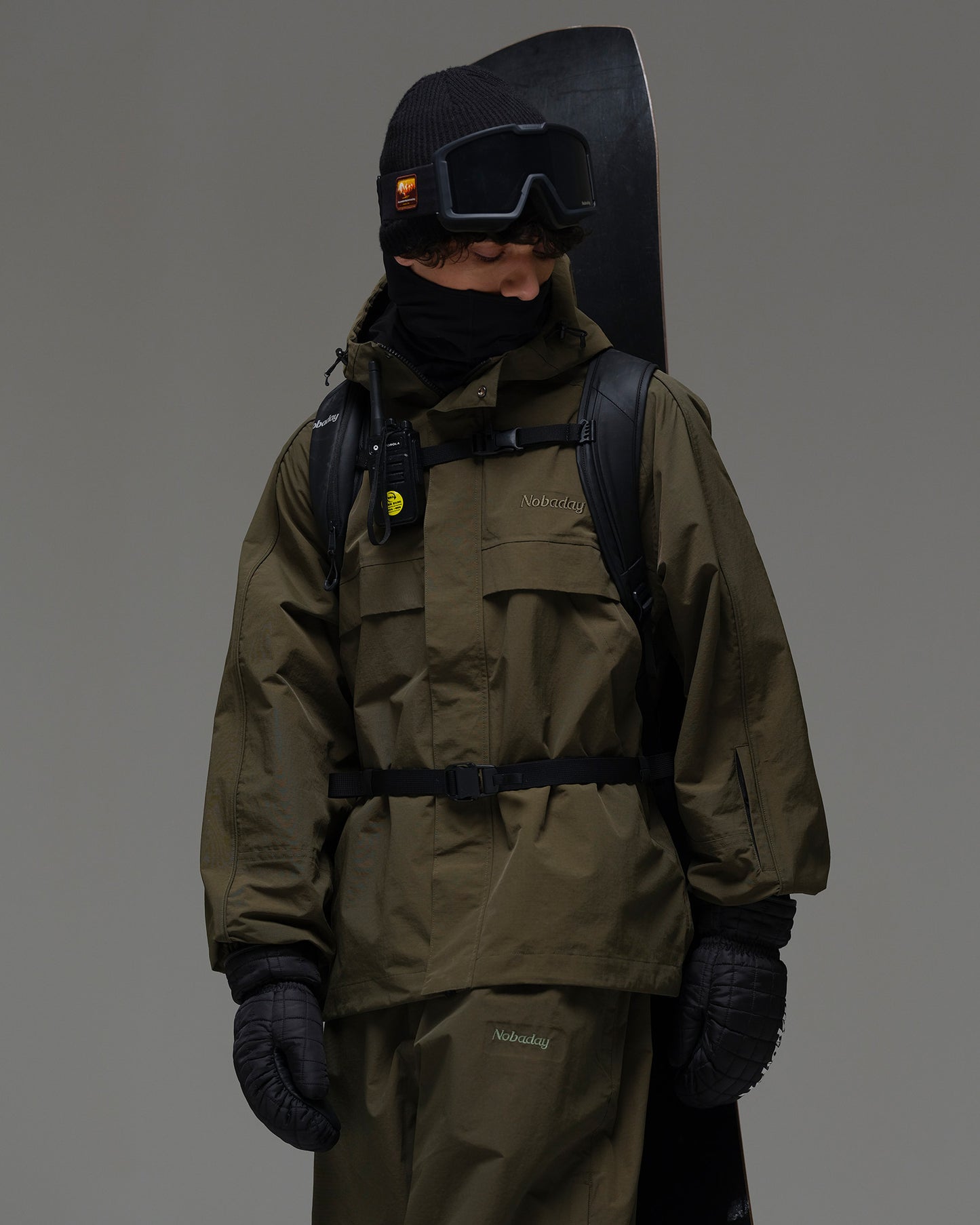 Man wearing a dark green NOBADAY snow jacket with a snowboard, backpack and winter gear