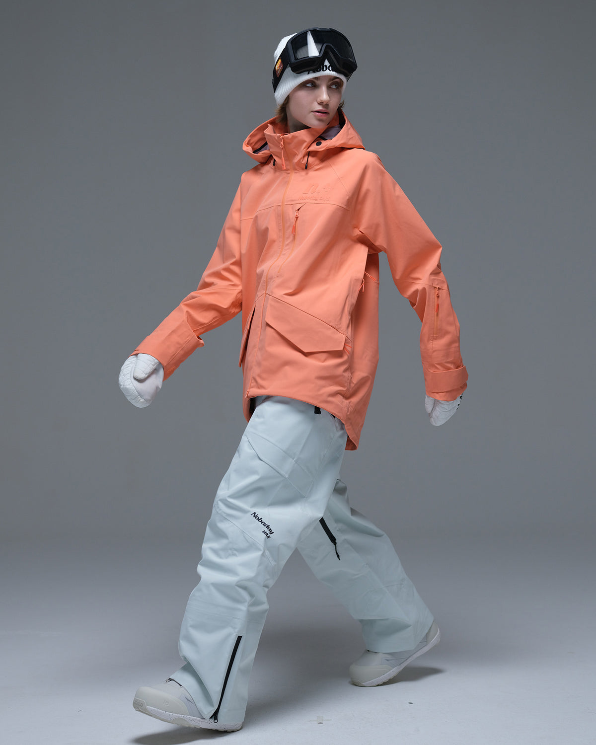 Full-body action view of a model wearing the coral orange Nobaday Women’s ArcticStorm Freeride Zip-Up 3L Snow Jacket with light blue snow pants