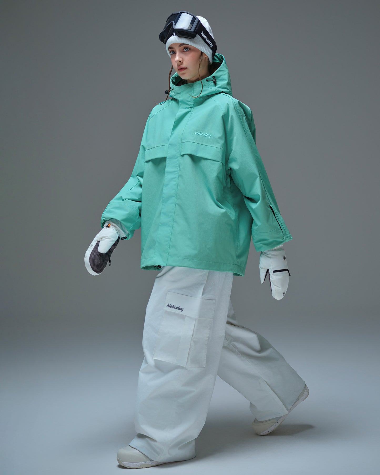 Full body side pose of Nobaday women's blue active snow jacket with white snow pants, mittens, oversized fit, and sleeve pass pocket