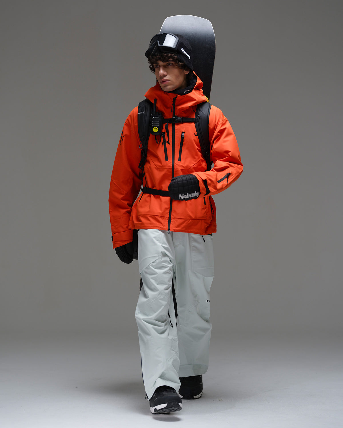 Full-body view of a male snowboarder wearing the NOBADAY Men’s MaxExtreme Pro 3L snow jacket in orange with a backpack and snowboard