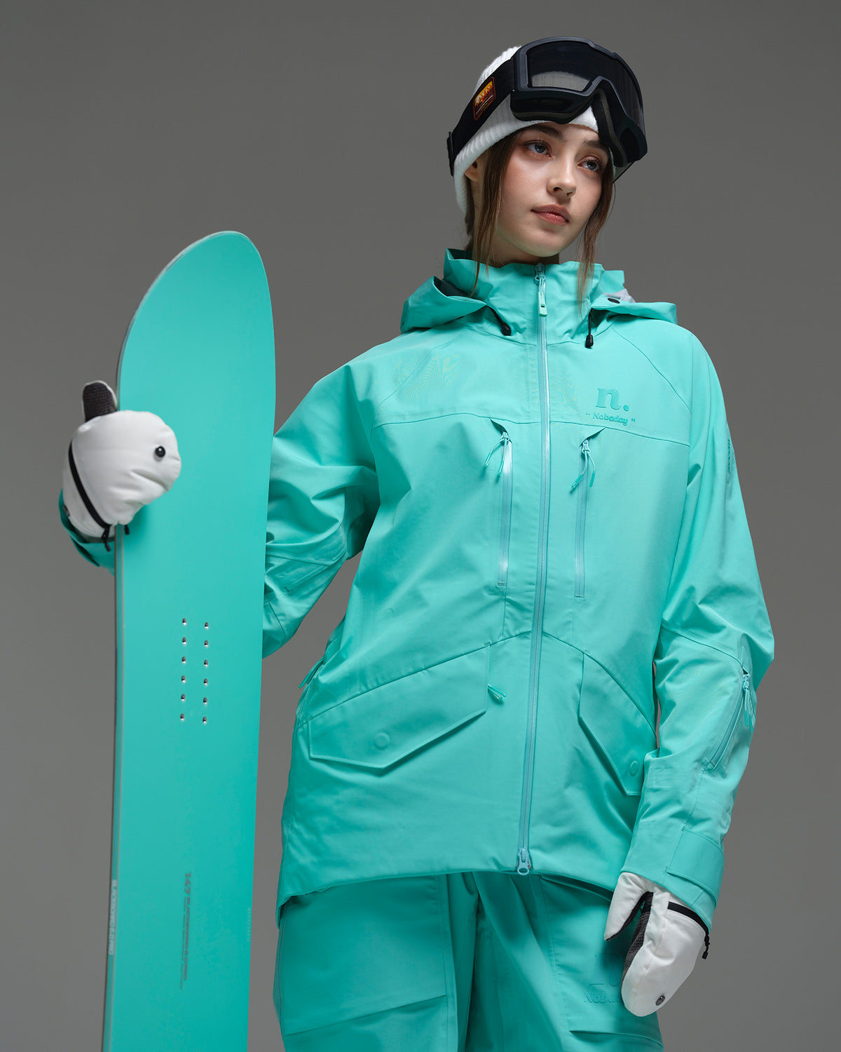 Woman wearing the NOBADAY Women's ArcticStorm Freeride 3L blue snow jacket while holding a matching snowboard