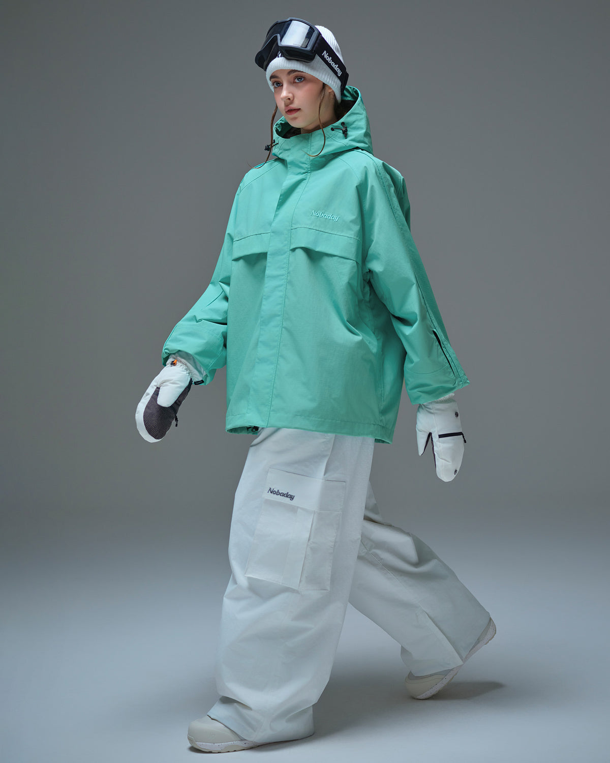 Full body side pose of Nobaday women's blue active snow jacket with white snow pants, mittens, oversized fit, and sleeve pass pocket