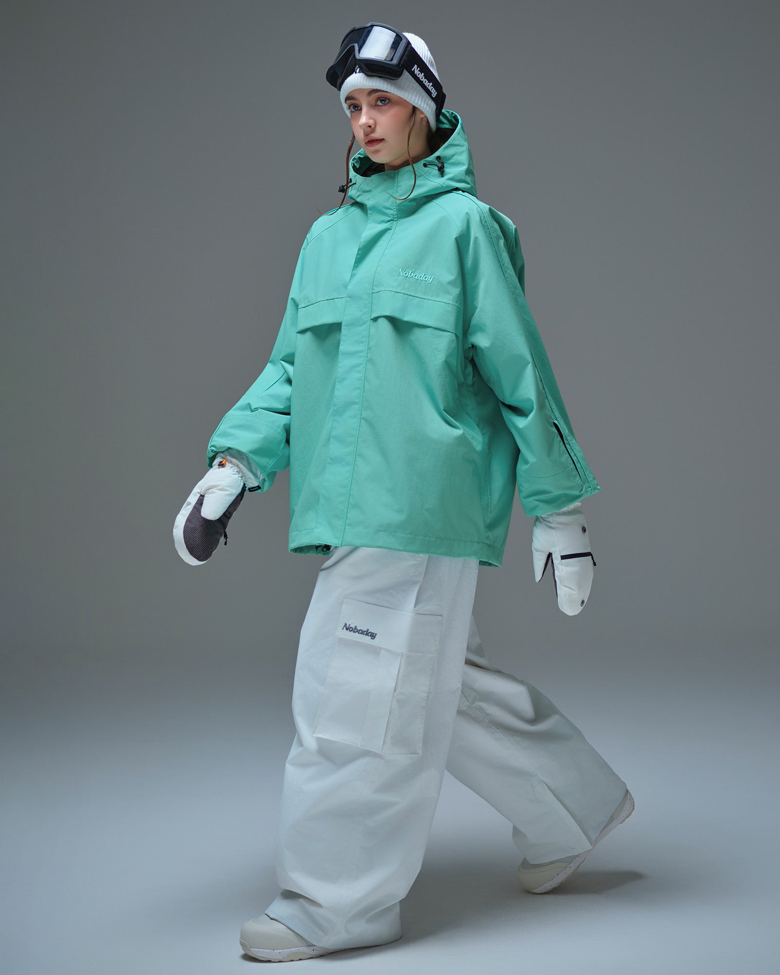 Full body side pose of Nobaday women's blue active snow jacket with white snow pants, mittens, oversized fit, and sleeve pass pocket