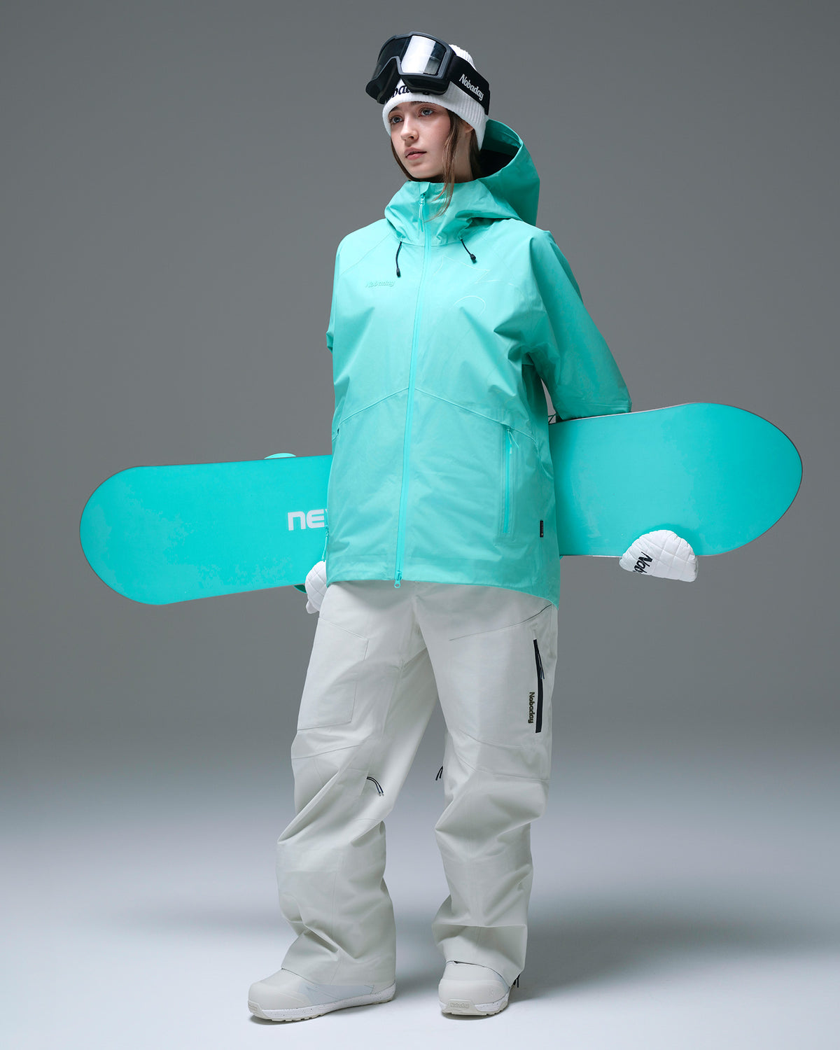 Woman wearing a Nobaday Blue GORE-TEX snowboard jacket with white snow pants and a snowboard