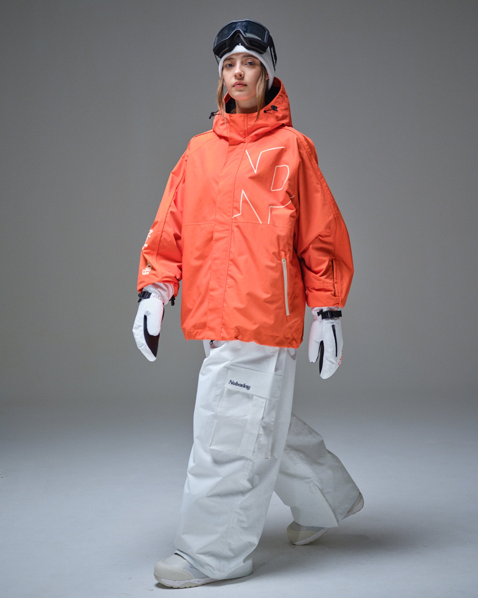 Full body side pose of Nobaday women's orange active snow jacket with white snow pants, mittens, oversized fit, and adjustable hood