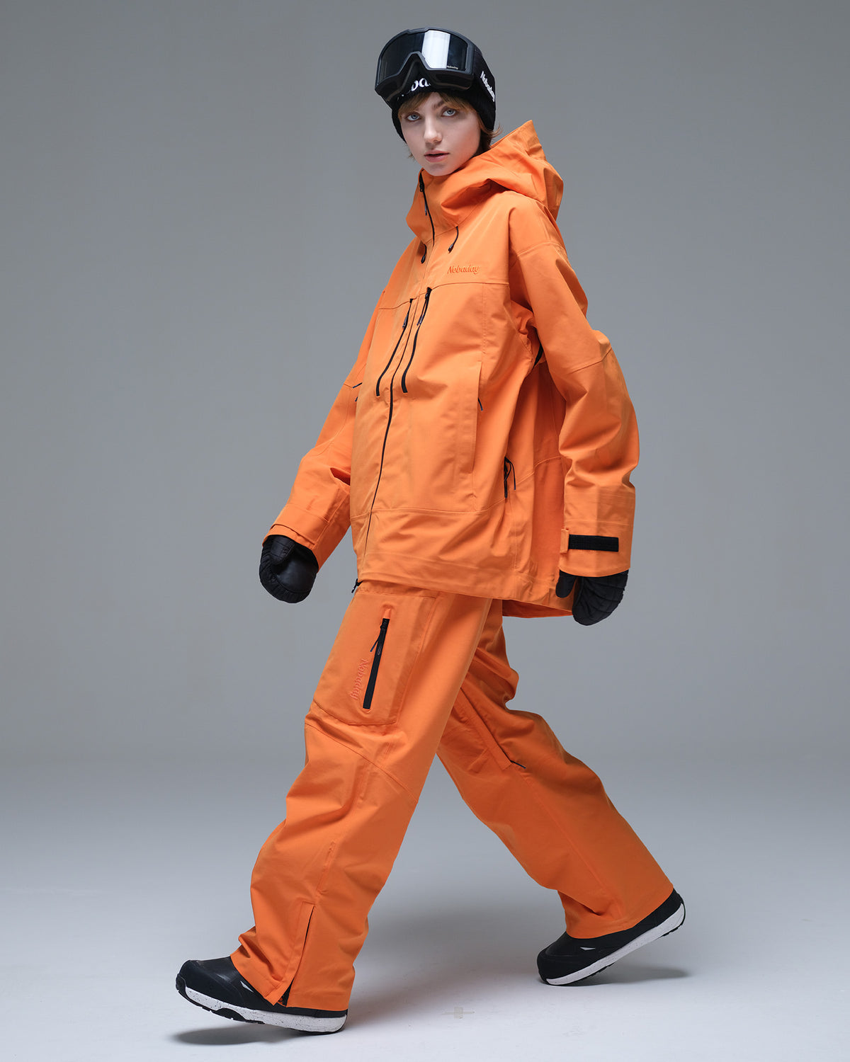 Full-body action pose of a woman wearing the NOBADAY Women's Pure Free 3L orange snow jacket with matching snow pants