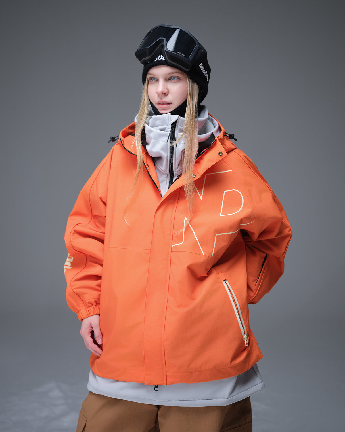 Nobaday Women's Pure Free Active orange snow jacket layered over a white midlayer with goggles, oversized sleeves, and waterproof shell