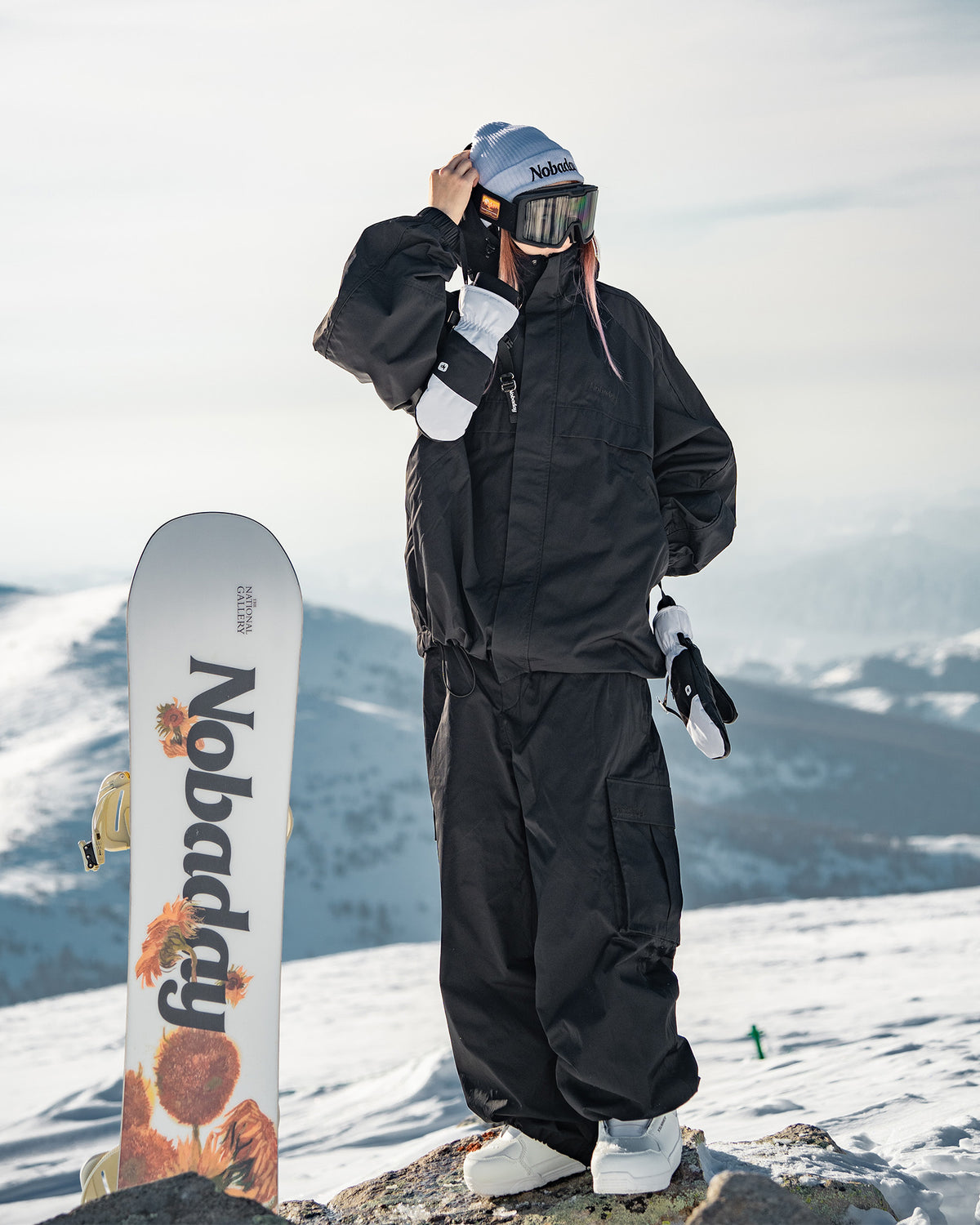Nobaday Women's Pure Free black snow jacket worn outdoors on snowy mountain with snowboard, goggles, mittens, and black snow pants