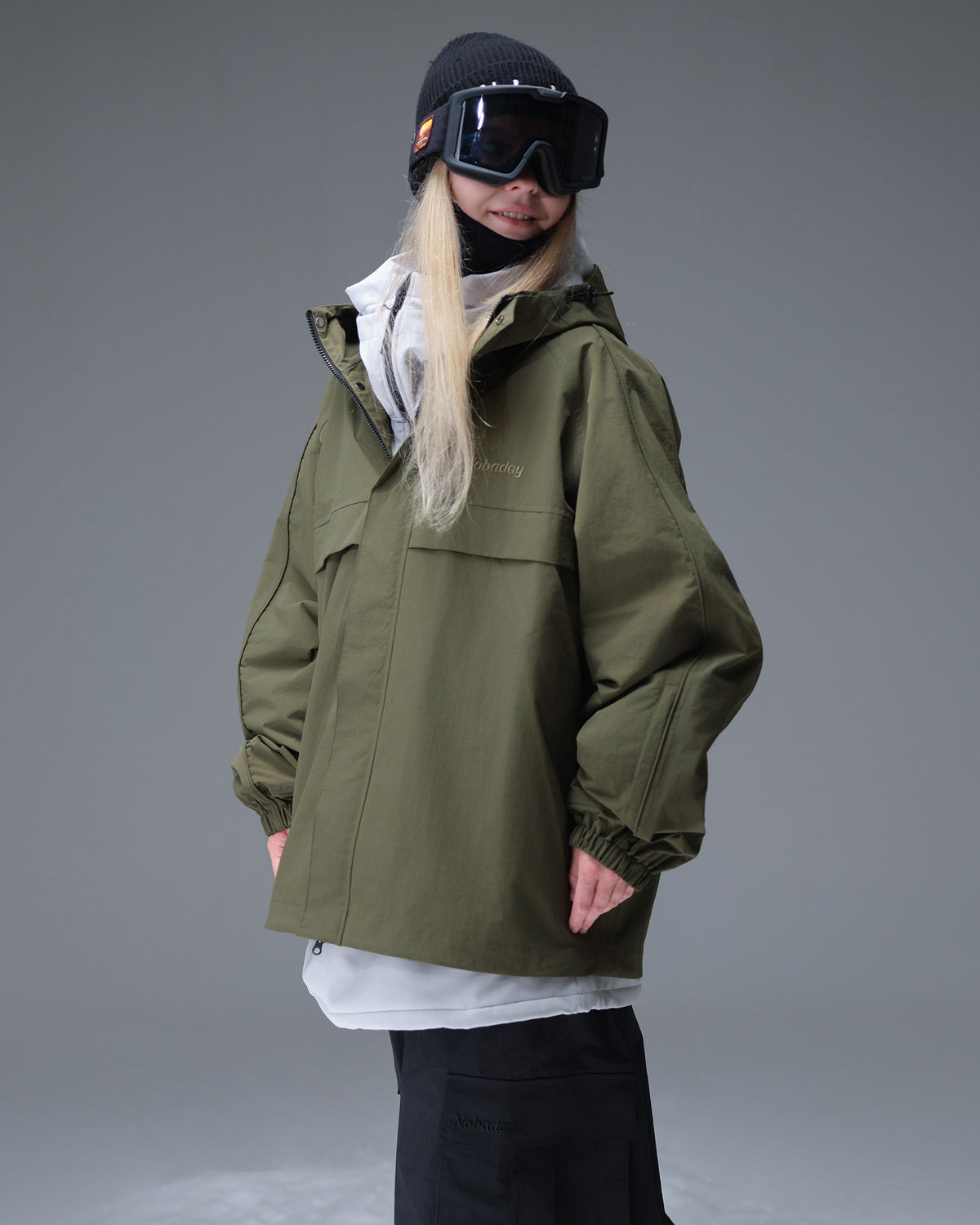 Nobaday Women's Pure Free Cargo green snow jacket layered over a white midlayer with goggles, roomy fit, and waterproof shell
